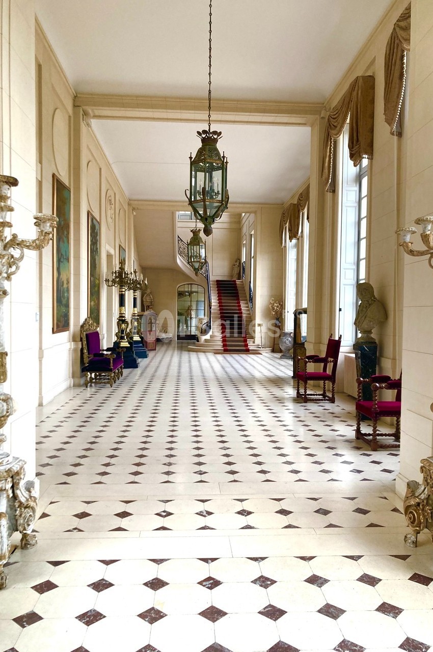 Hall spacieux avec sol en damier, lustres anciens, chaises décoratives et escalier au fond.