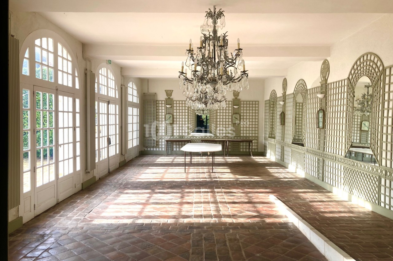 Salle lumineuse avec sol en carreaux de terre cuite, grandes fenêtres, miroirs muraux et lustre central en cristal.