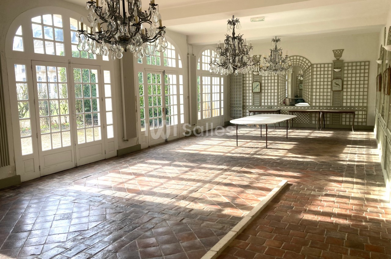 Salle lumineuse avec sol en carreaux de terre cuite, grandes fenêtres cintrées, miroirs et lustres en cristal.