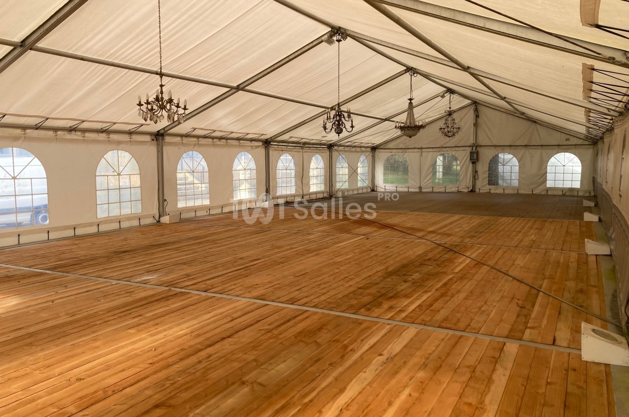Intérieur d'une grande tente avec un sol en bois, des lustres suspendus et des fenêtres en arc sur les côtés.