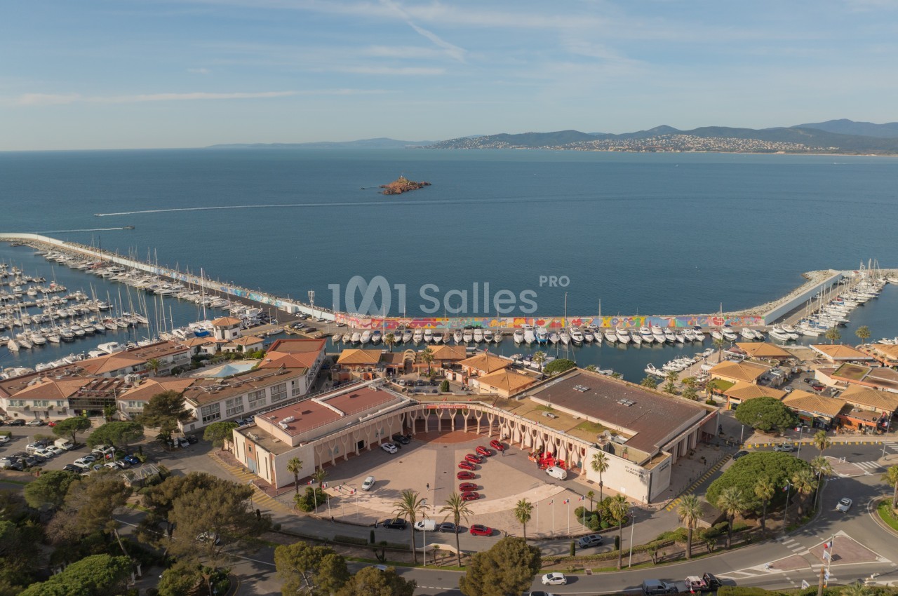 Vue aérienne d'un port de plaisance avec des voiliers alignés, une esplanade et la mer en arrière-plan.