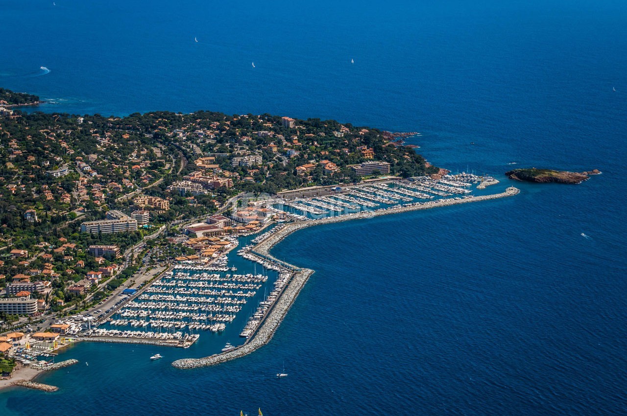 Vue aérienne d'un port de plaisance entouré de bâtiments résidentiels et de végétation, bordé par la mer.