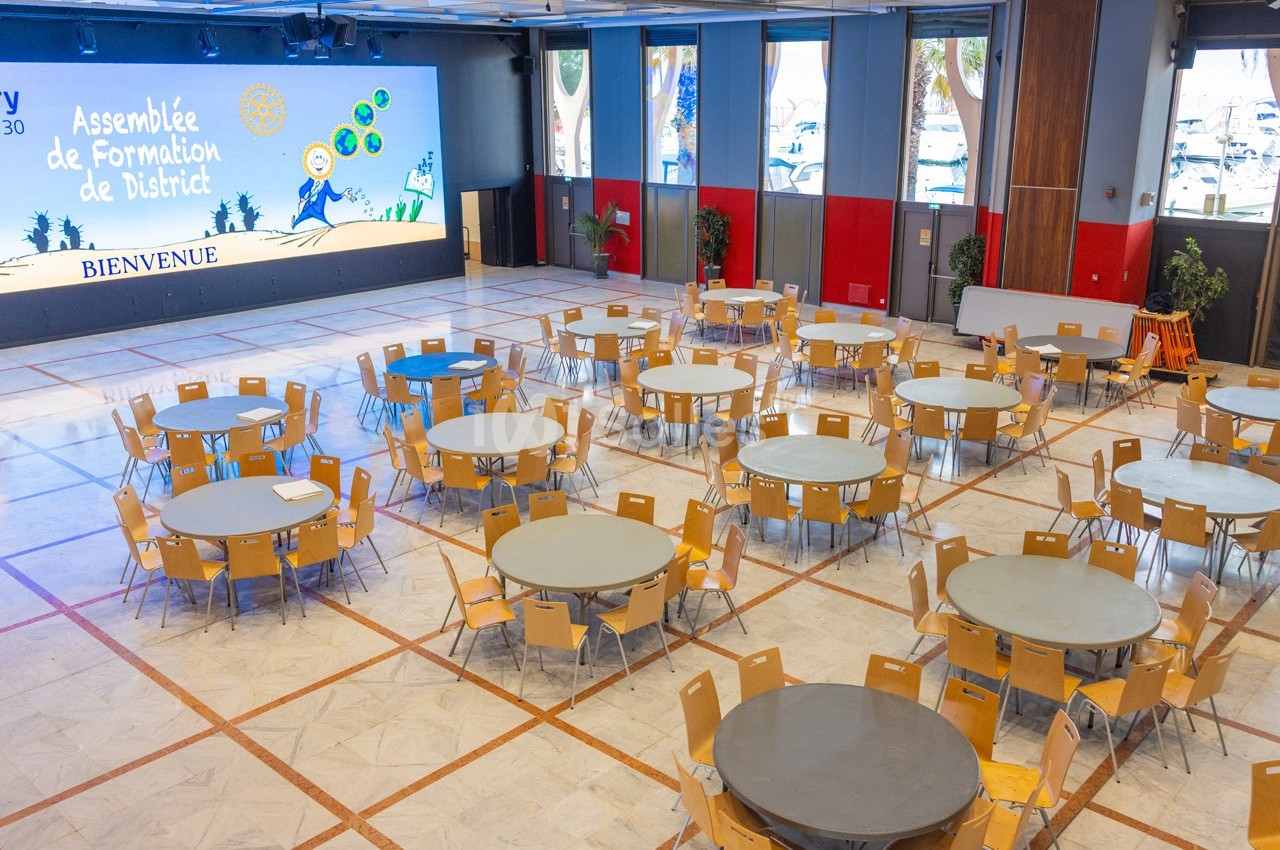 Salle de conférence avec des tables rondes et des chaises disposées, écran de présentation en arrière-plan.