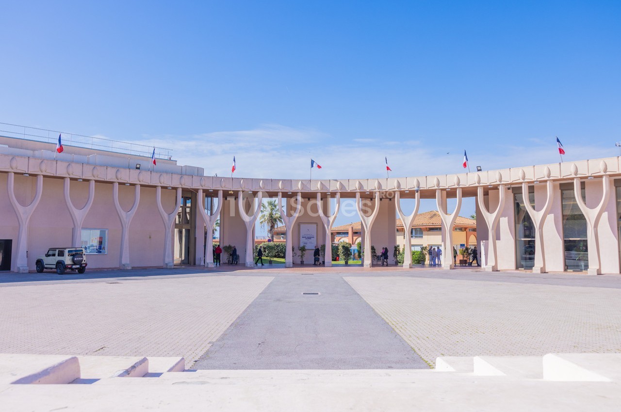 Vue d'une place pavée entourée d'un bâtiment moderne avec des colonnes en forme d'arbres et des drapeaux français.