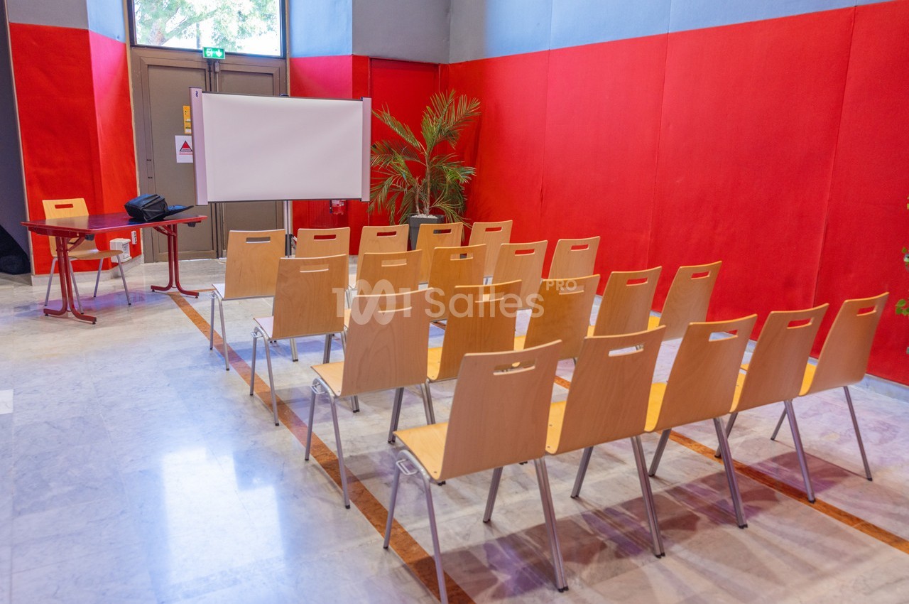Salle de réunion avec des chaises en bois alignées face à un écran blanc, entourée de murs rouges.