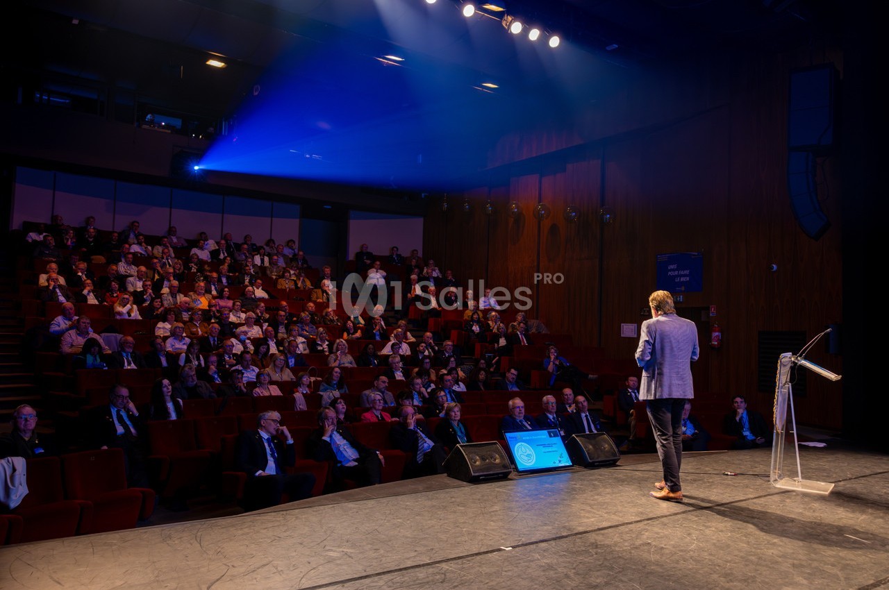 Un conférencier s'adresse à un public assis dans une salle de spectacle éclairée par des projecteurs.