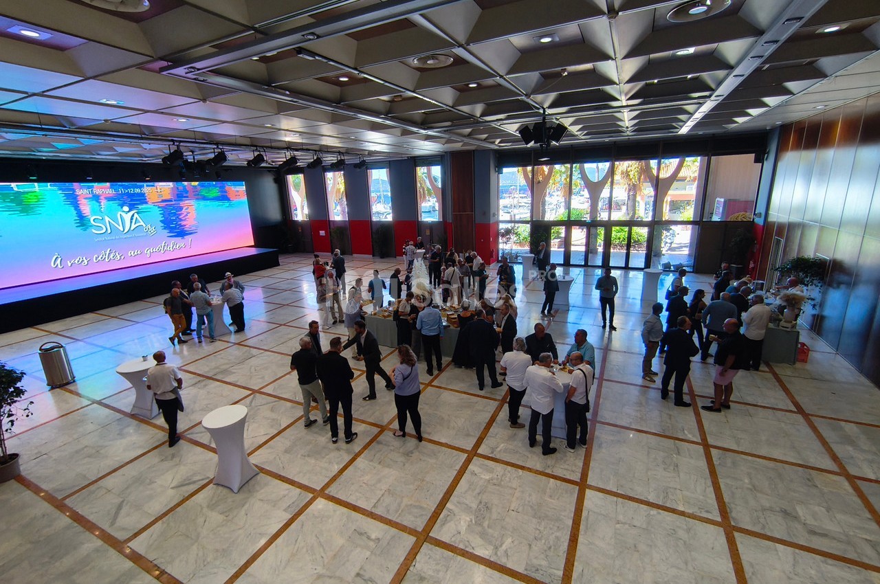 Groupe de personnes rassemblées dans une grande salle lumineuse avec un écran géant et des baies vitrées en arrière-plan.