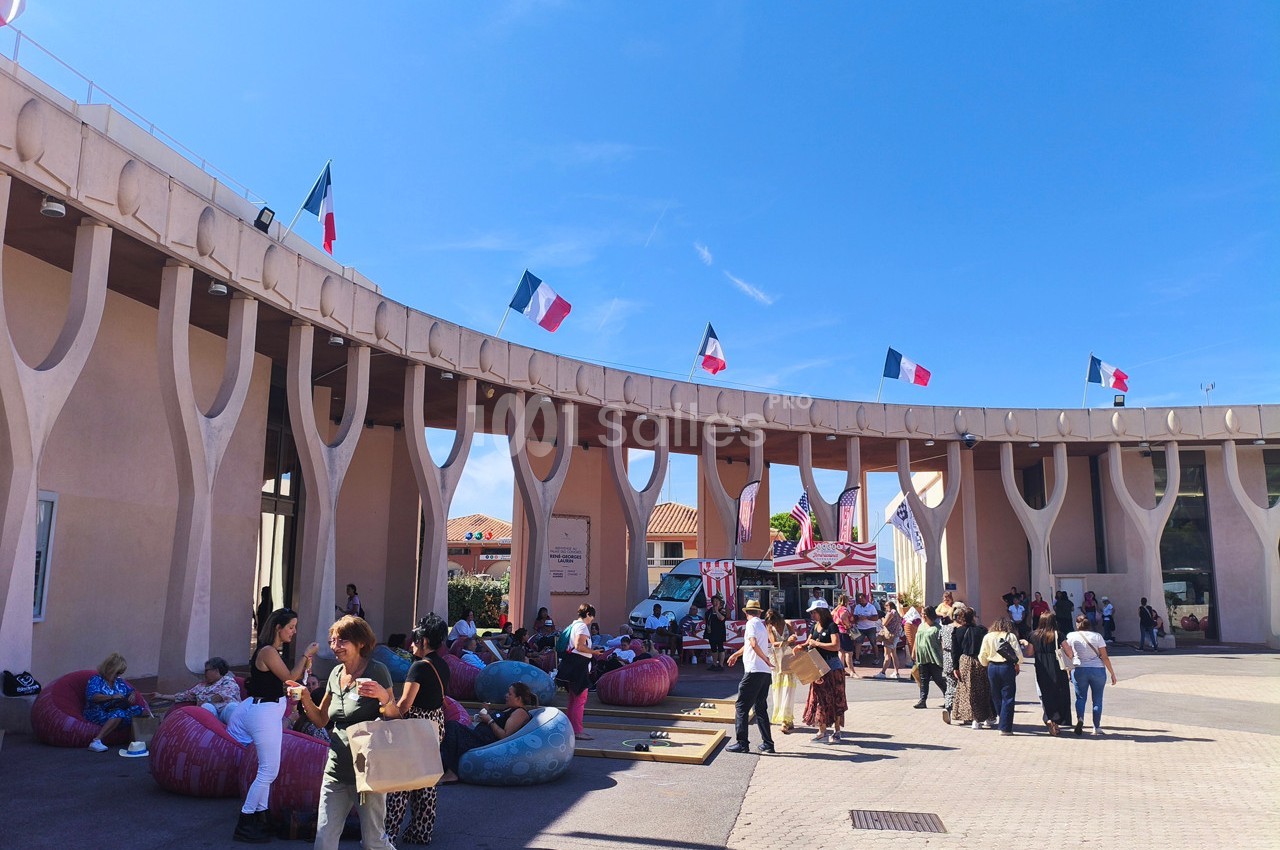 Des personnes se rassemblent devant un bâtiment moderne orné de drapeaux français, sous un ciel dégagé.