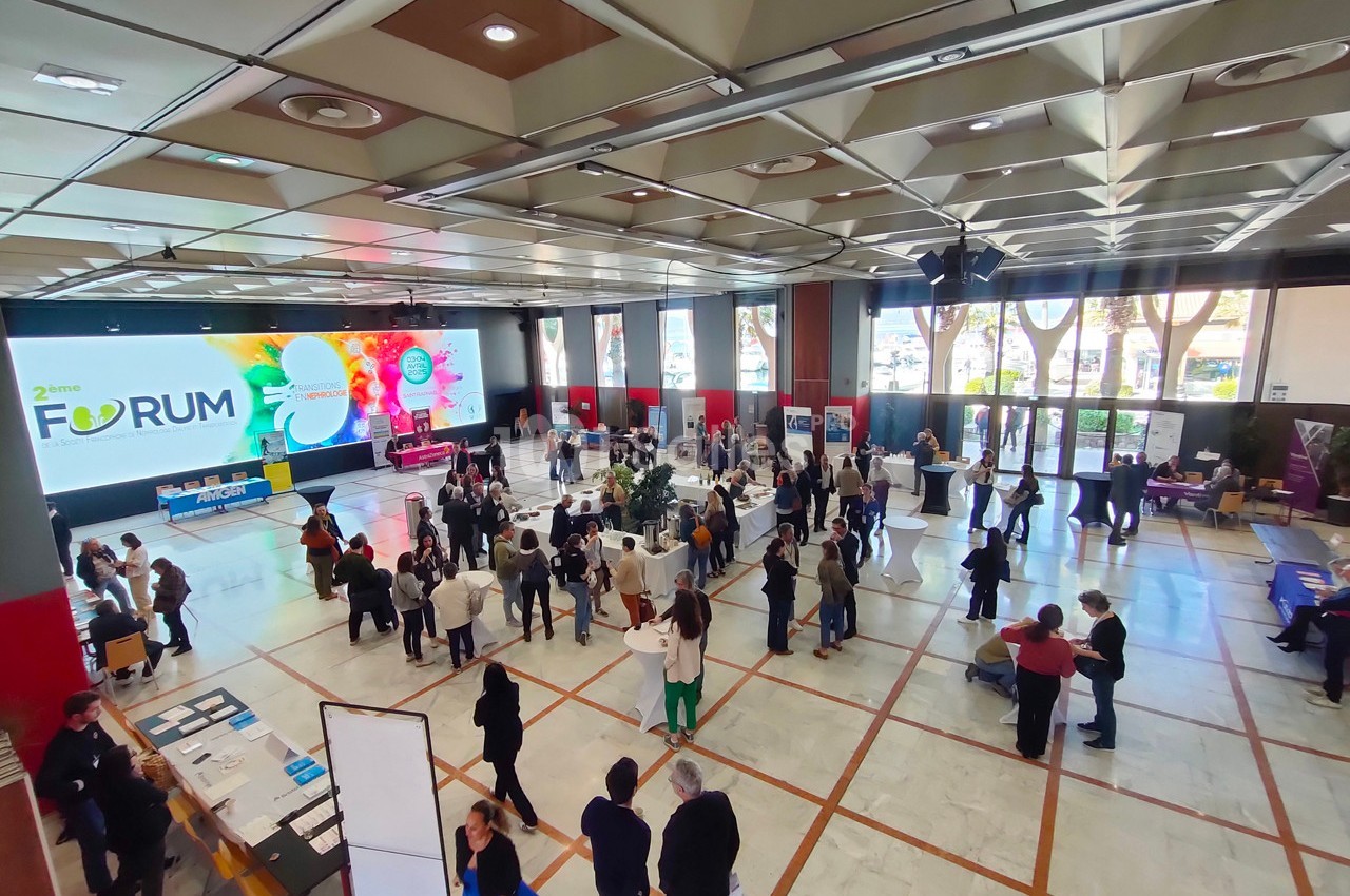 Vue d'ensemble d'un forum professionnel avec des stands, des participants en discussion et un écran affichant le logo de l…