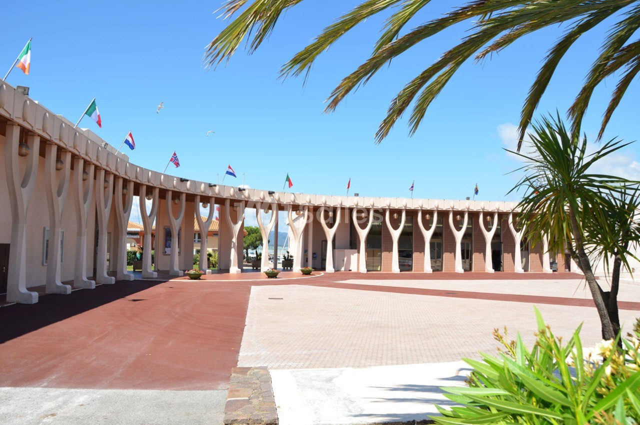 Bâtiment moderne en forme de demi-cercle avec des drapeaux internationaux, entouré de palmiers sous un ciel bleu.