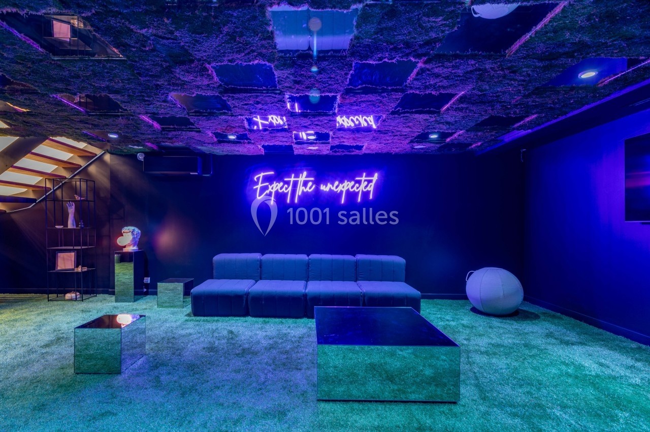 Salle moderne avec canapé gris, tables basses réfléchissantes, plafond végétalisé et néon ’Expect the unexpected’.