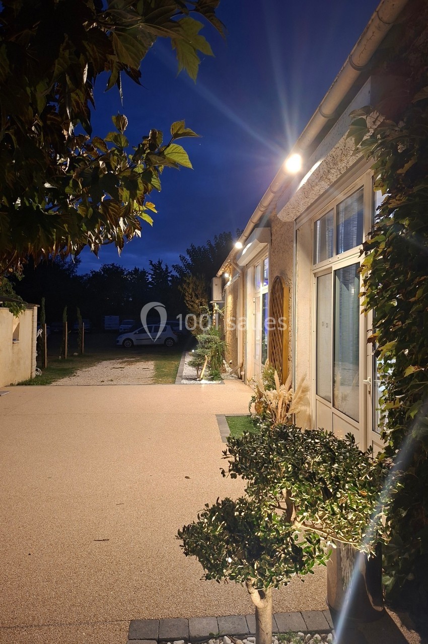 Cour extérieure éclairée en soirée, bordée de bâtiments avec des plantes et un arbre au premier plan.