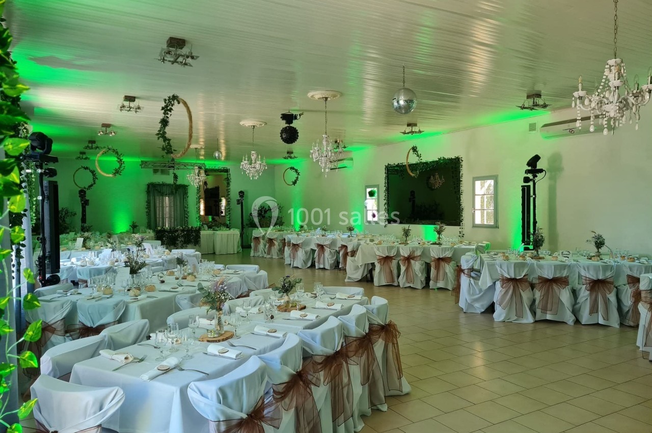 Salle de réception décorée avec des tables dressées, nappes blanches, chaises ornées de nœuds marron et éclairage vert.