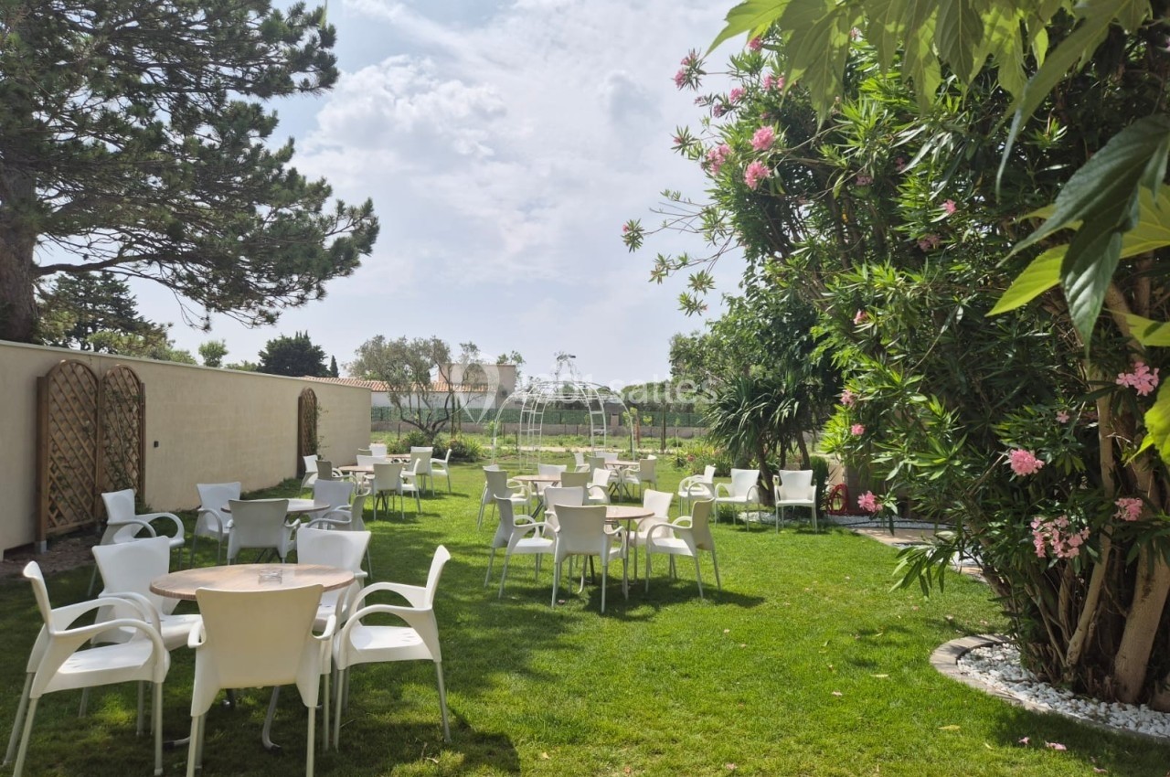 Jardin avec tables et chaises blanches disposées sur une pelouse, entouré d'arbres et de fleurs sous un ciel dégagé.