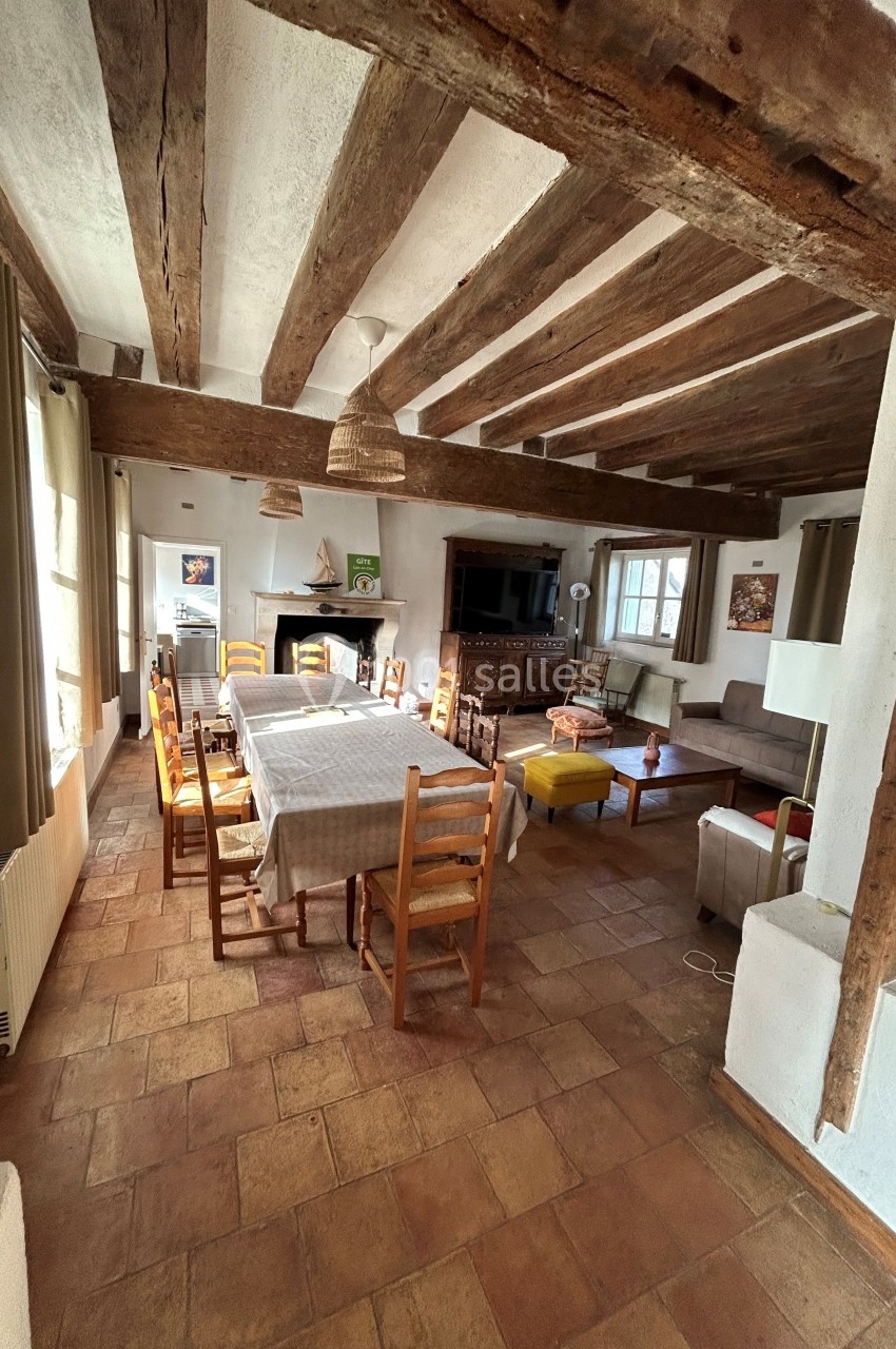 Salle à manger rustique avec poutres en bois, table dressée, chaises en bois et coin salon avec canapés et cheminée.