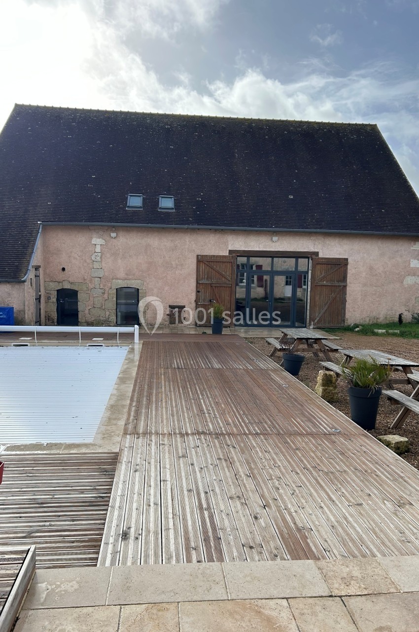 Grange rénovée avec façade en pierre, terrasse en bois et piscine partiellement couverte au premier plan.