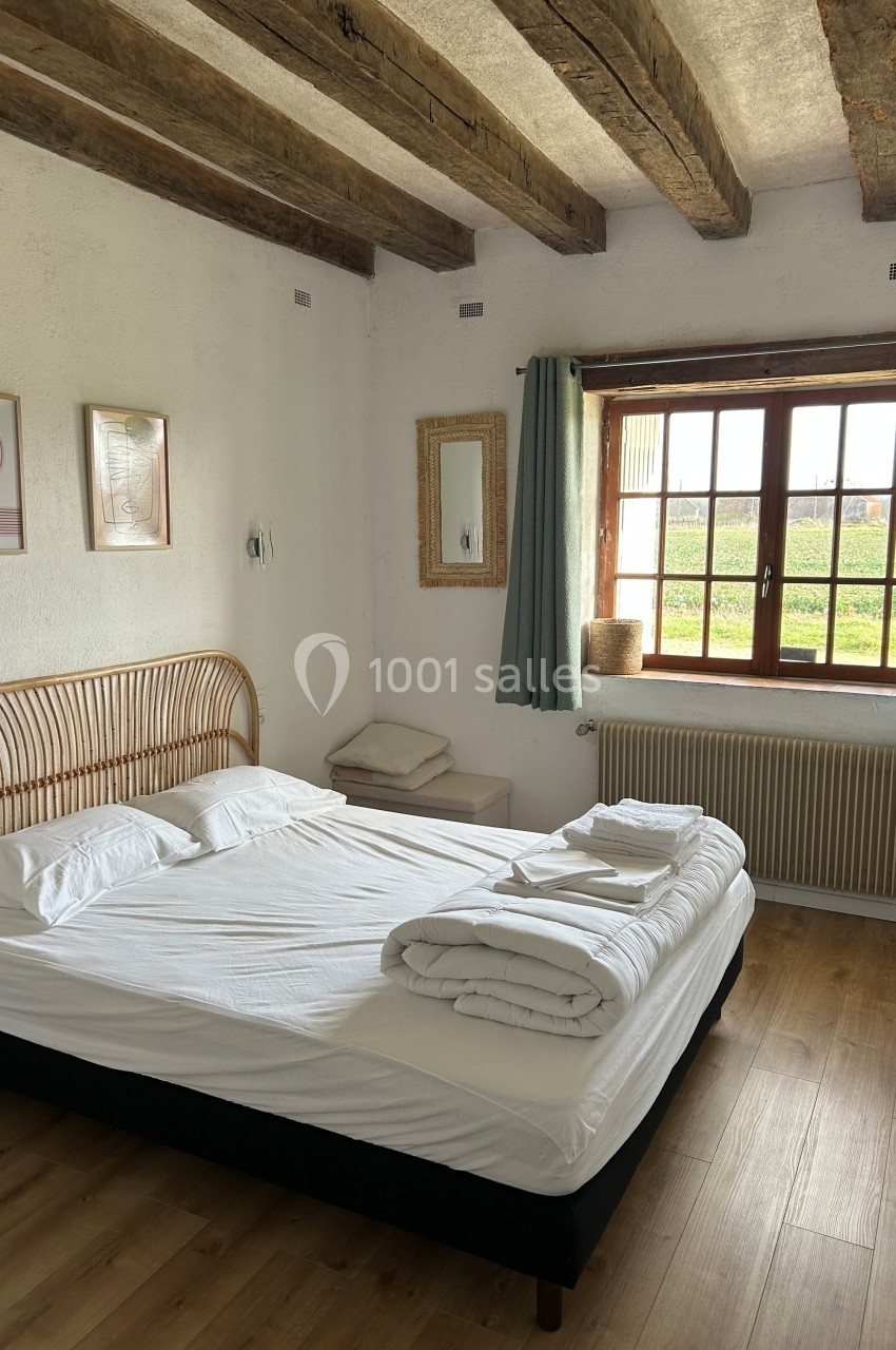 Chambre lumineuse avec lit double, poutres apparentes, fenêtre donnant sur un paysage verdoyant et décoration minimaliste.