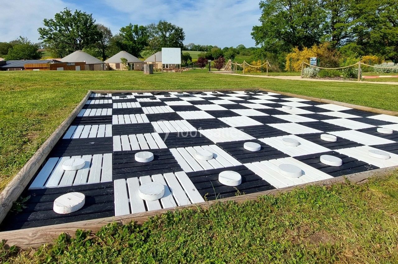 Jeu de dames géant en plein air avec des pions blancs sur un damier noir et blanc, entouré de pelouse.