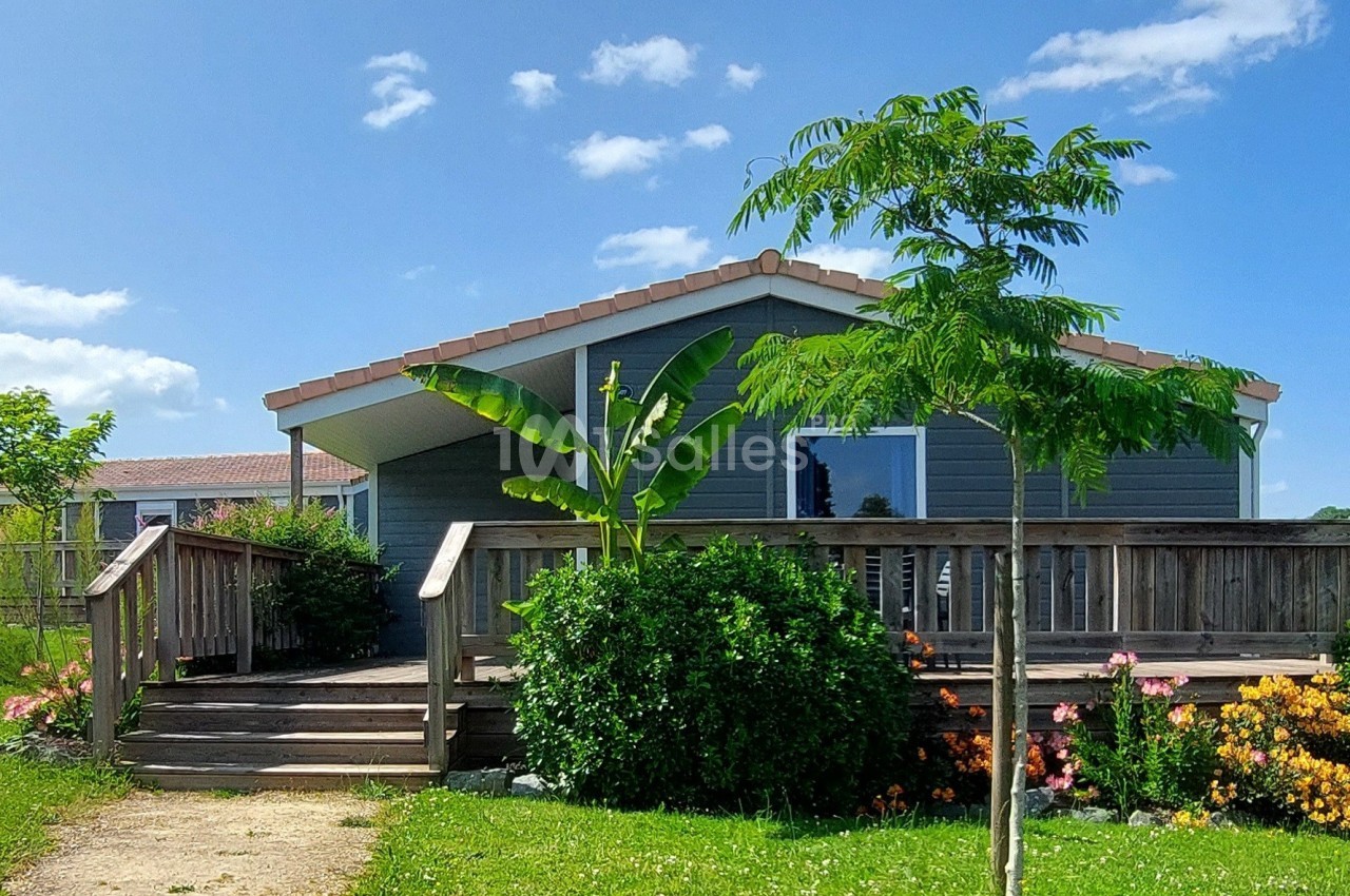 Maison en bois avec terrasse entourée de végétation, dont un bananier et des fleurs colorées, sous un ciel bleu.