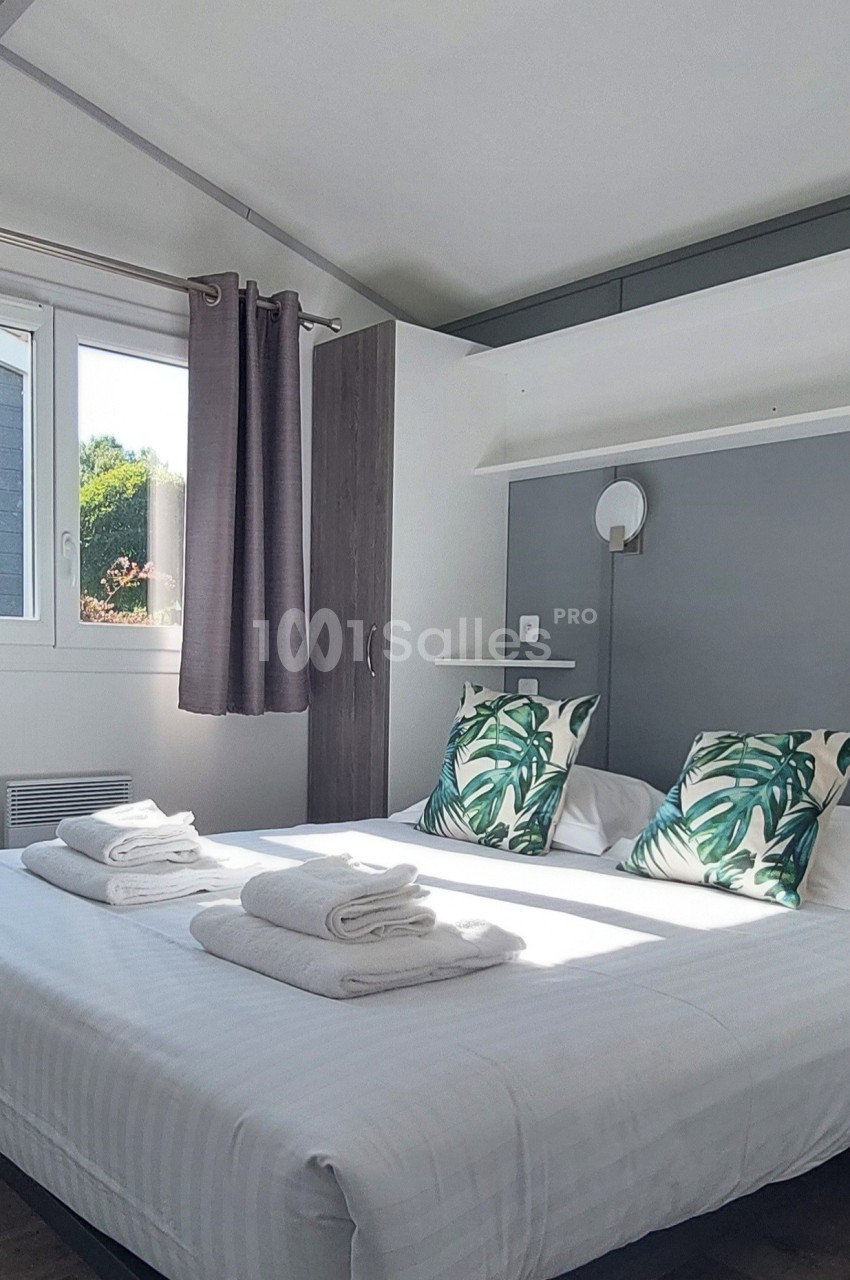Chambre lumineuse avec un lit double, draps blancs, coussins à motifs végétaux, et fenêtre avec rideaux violets.