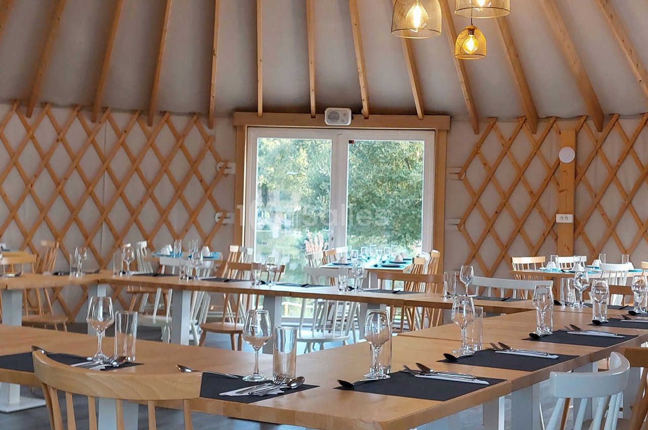 Salle de restaurant lumineuse avec tables en bois dressées, chaises assorties et grande baie vitrée donnant sur l'extérieur.