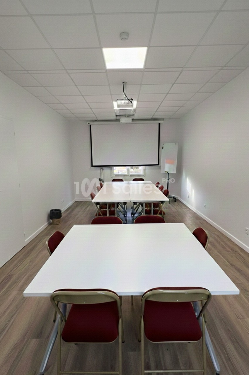 Salle de réunion lumineuse avec tables blanches, chaises rouges, projecteur, écran et tableau blanc.