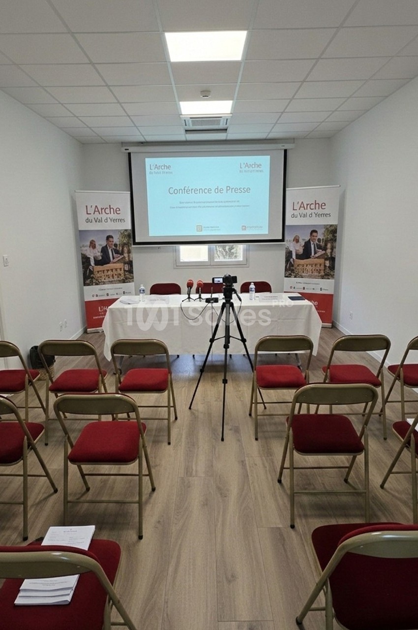 Salle de conférence avec chaises alignées, table équipée de micros et écran affichant ’Conférence de Presse’.