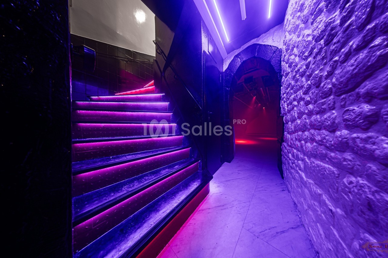 Escalier éclairé par des néons violets menant à un couloir en pierre dans une ambiance tamisée.