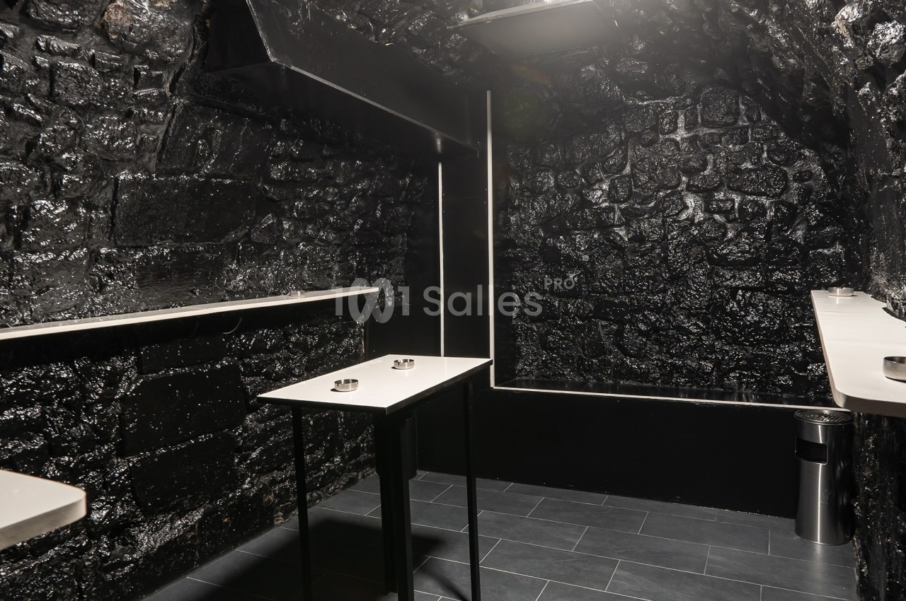 Salle sombre avec murs en pierre noire, tables hautes blanches et sol en carrelage gris.