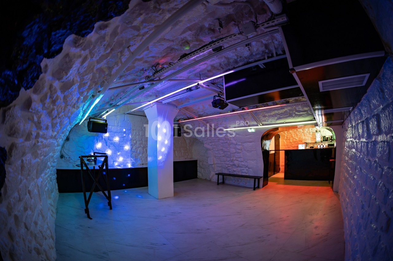 Salle voûtée en pierre blanche avec éclairage coloré, piste de danse vide et comptoir de bar au fond.