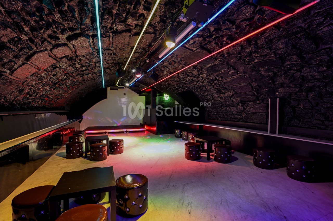 Salle voûtée en pierre avec éclairage néon coloré, tables basses et tabourets disposés sur un sol clair.