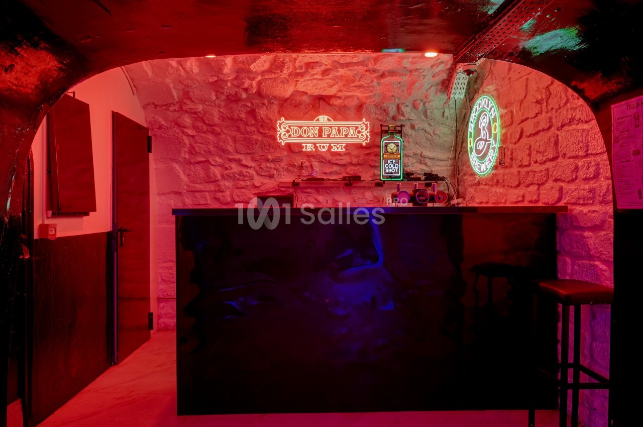 Bar avec comptoir noir, éclairé par des néons rouges et verts, dans une pièce aux murs en pierre apparente.