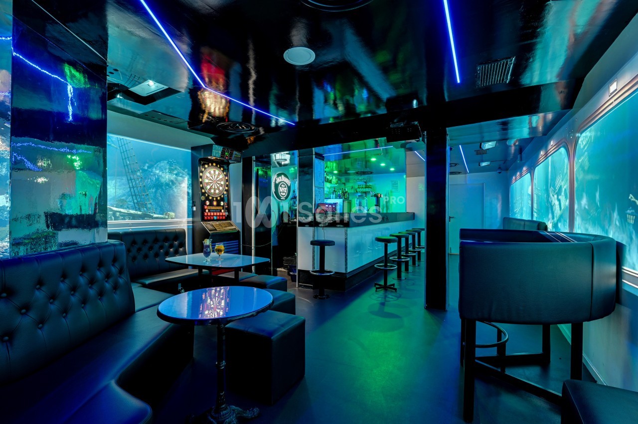 Intérieur d'un bar moderne avec éclairage néon, banquettes noires, tables basses et un jeu de fléchettes au mur.