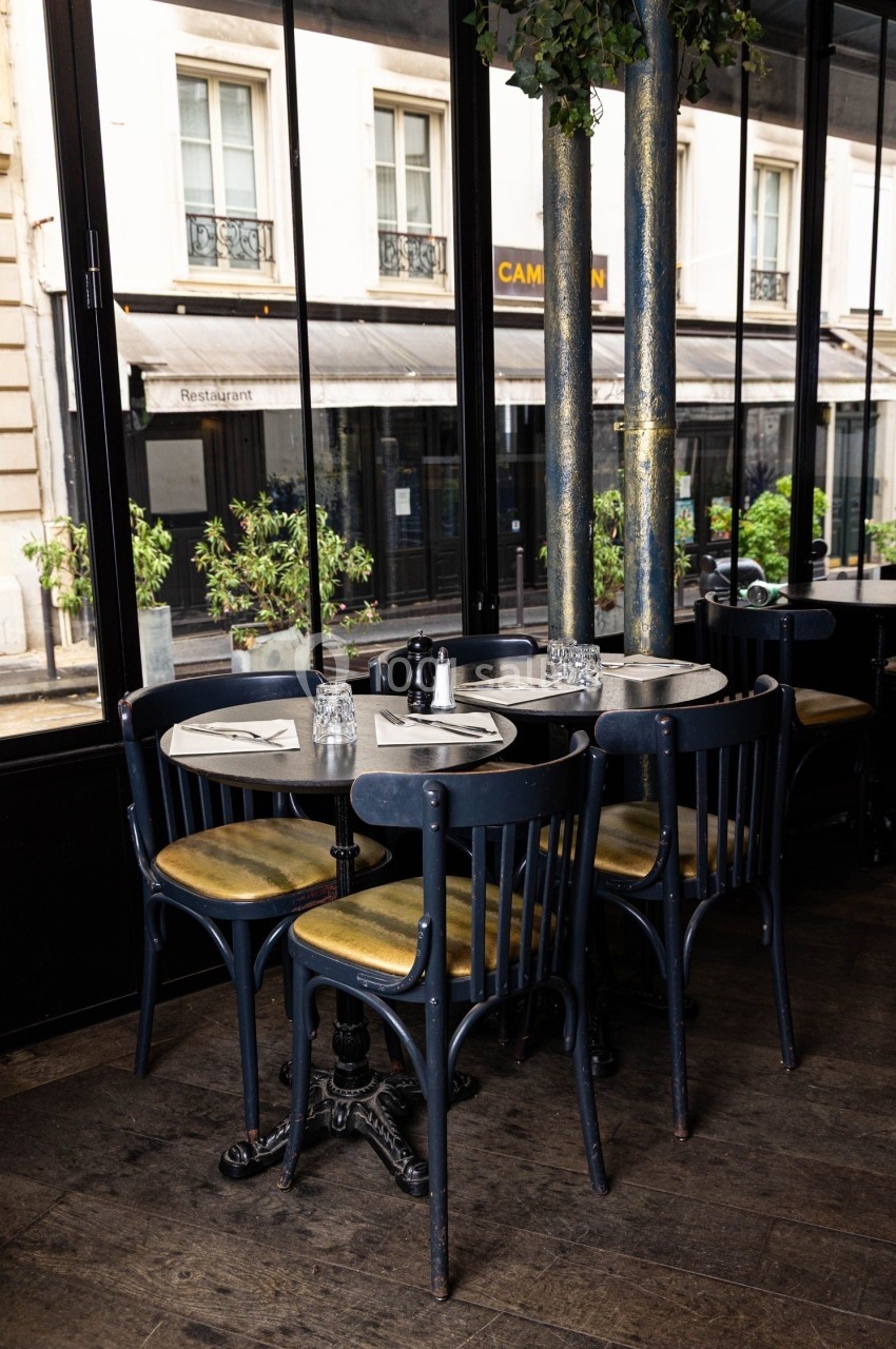 Tables rondes avec chaises en bois noir dans un café lumineux, avec vue sur des vitrines et des plantes extérieures.
