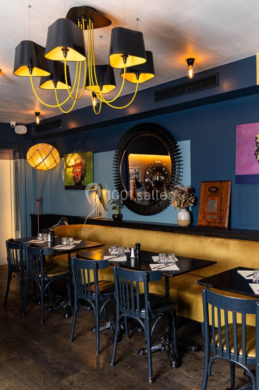 Salle de restaurant avec murs bleus, banquette jaune, tables noires, miroir rond et lustre moderne noir et jaune.