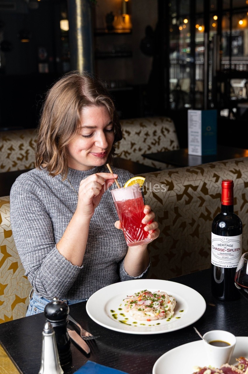 Une femme boit un cocktail rose avec une paille, assise à une table de restaurant avec un plat et une bouteille de vin.
