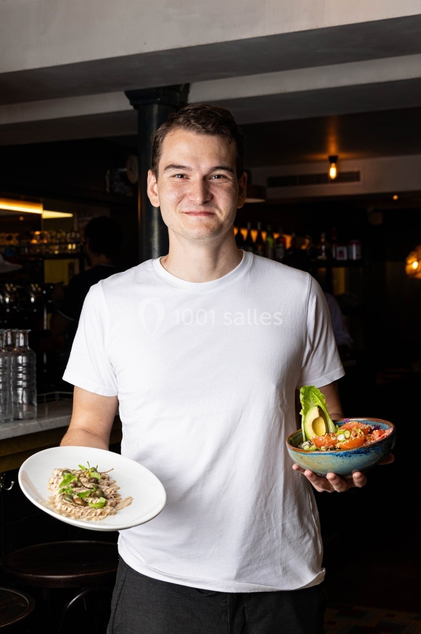 Un homme souriant tient un plat de risotto et un bol de salade garnie d'avocat dans un restaurant.