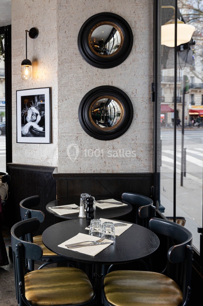 Salle de restaurant avec tables dressées, miroirs ronds au mur et vue sur une rue parisienne à travers une baie vitrée.
