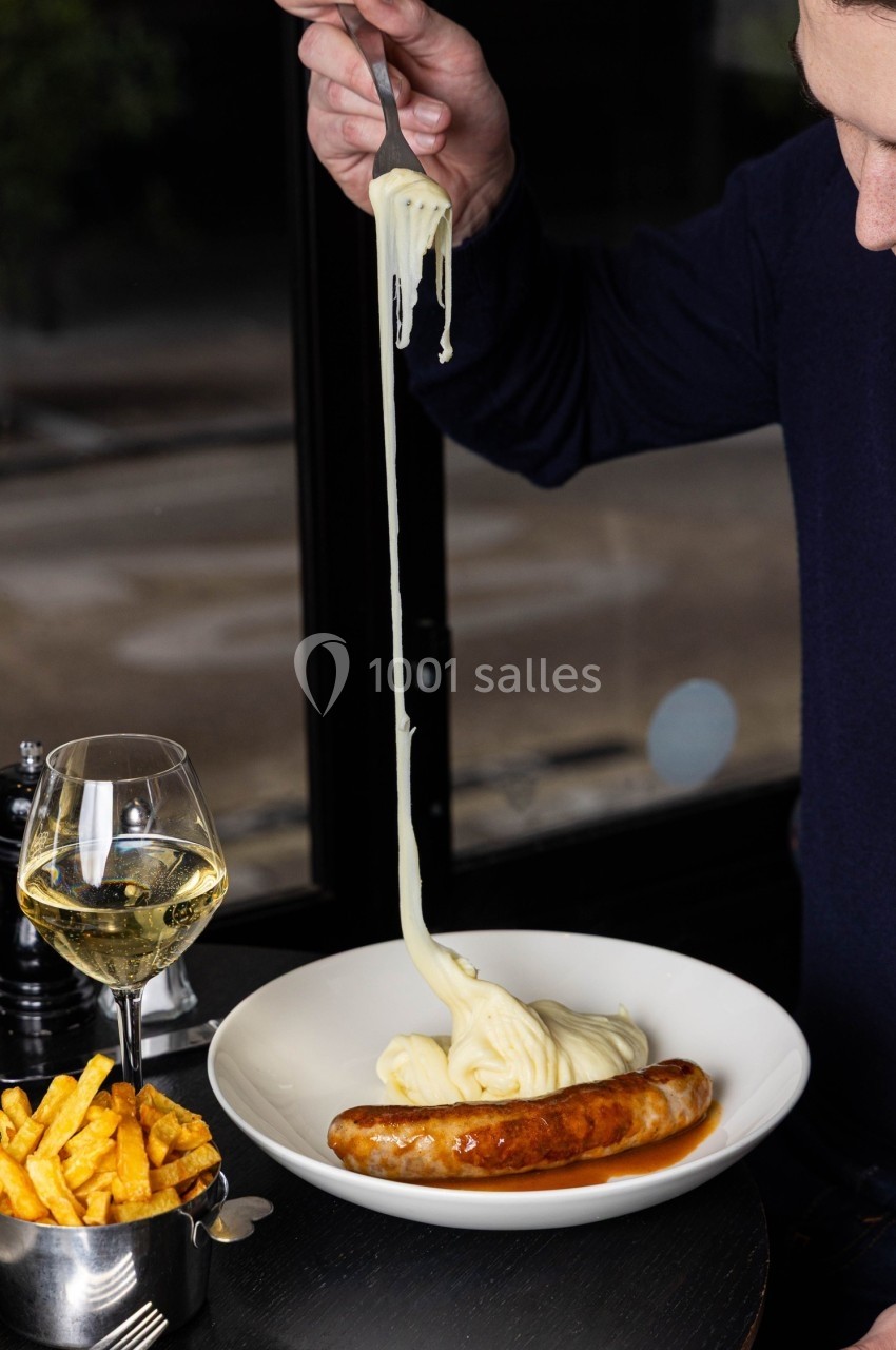 Une assiette contenant une saucisse, de la purée filante et des frites, avec un verre de vin blanc à côté.