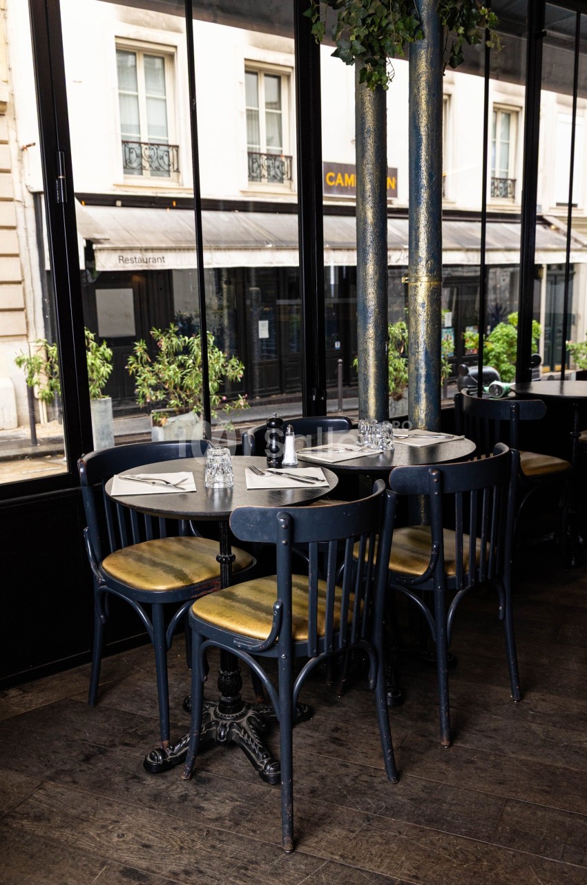Tables rondes avec chaises en bois noir dans un café lumineux, donnant sur une rue bordée de vitrines et de plantes.