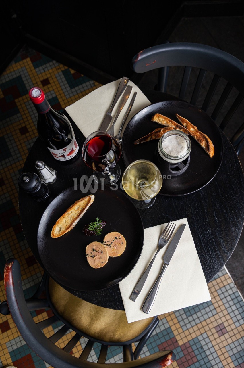 Table ronde avec assiettes de foie gras, pain grillé, verres de vin et bière, sur un sol en mosaïque colorée.