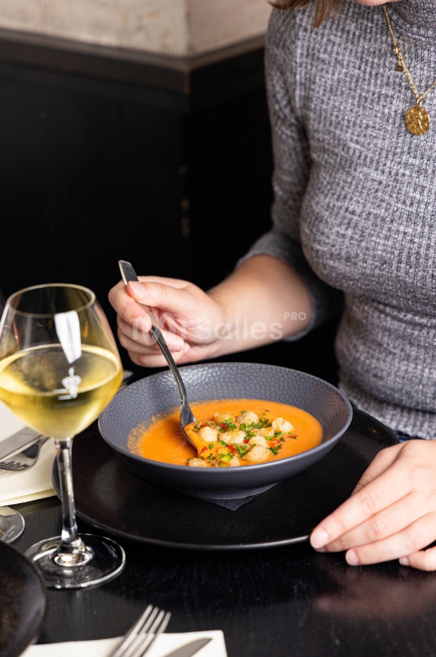 Une personne déguste une soupe orange garnie de croûtons et d'herbes, avec un verre de vin blanc à côté.