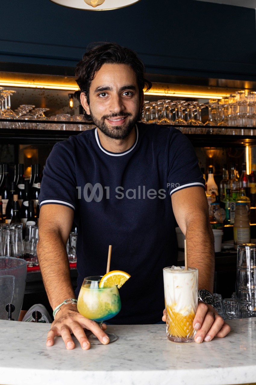 Un barman souriant présente deux cocktails colorés sur un comptoir en marbre dans un bar élégant.
