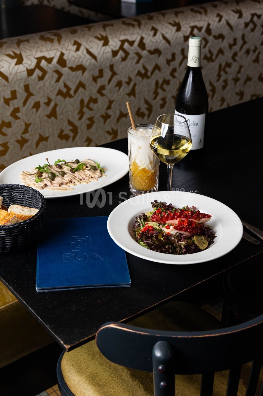 Table dressée avec des plats raffinés, une bouteille de vin, un verre de vin blanc et un cocktail, dans un cadre élégant.