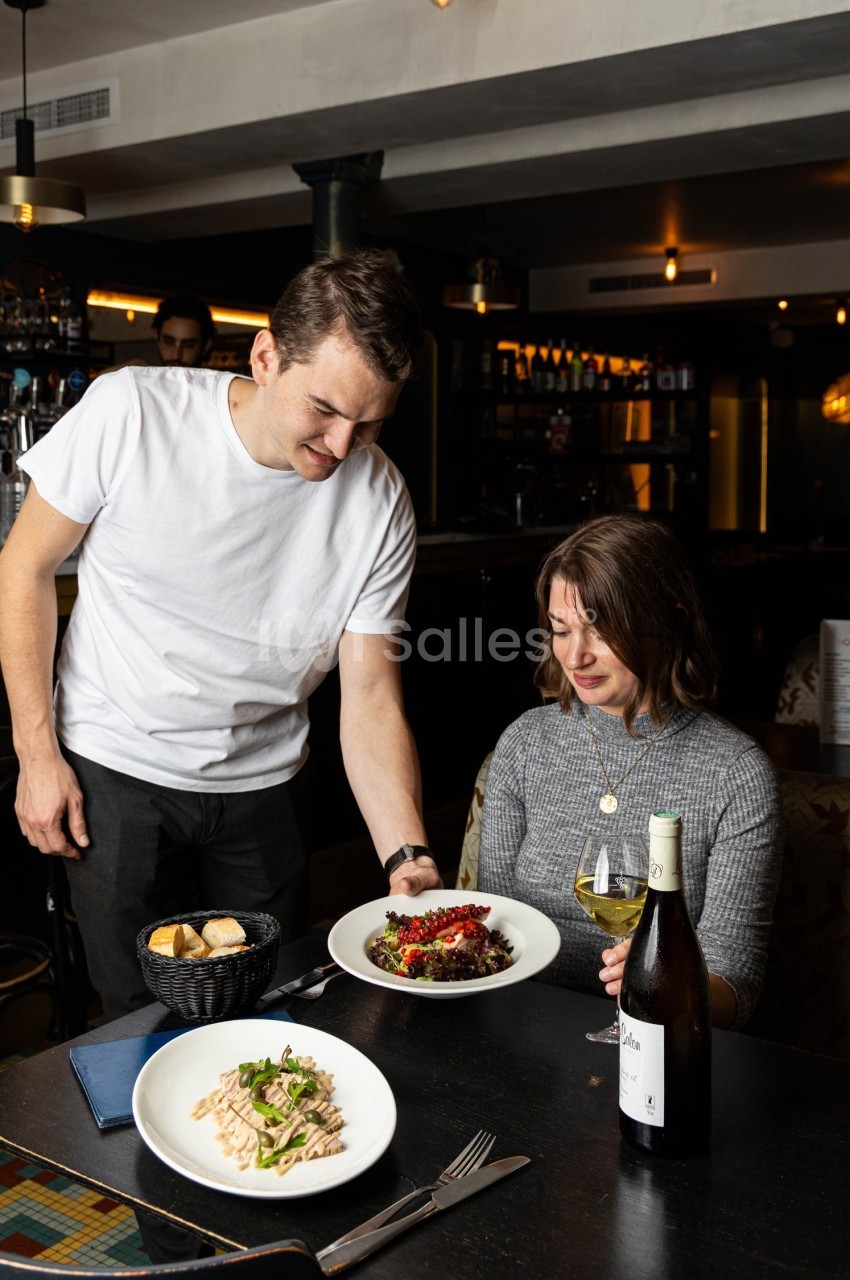 Un serveur sert un plat à une cliente assise dans un restaurant, avec des plats et une bouteille de vin sur la table.