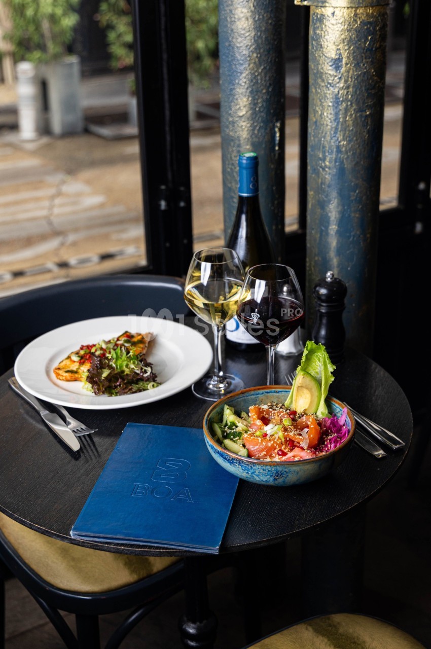 Table avec un bol de poke, une assiette de salade, deux verres de vin et une bouteille, dans un cadre élégant.