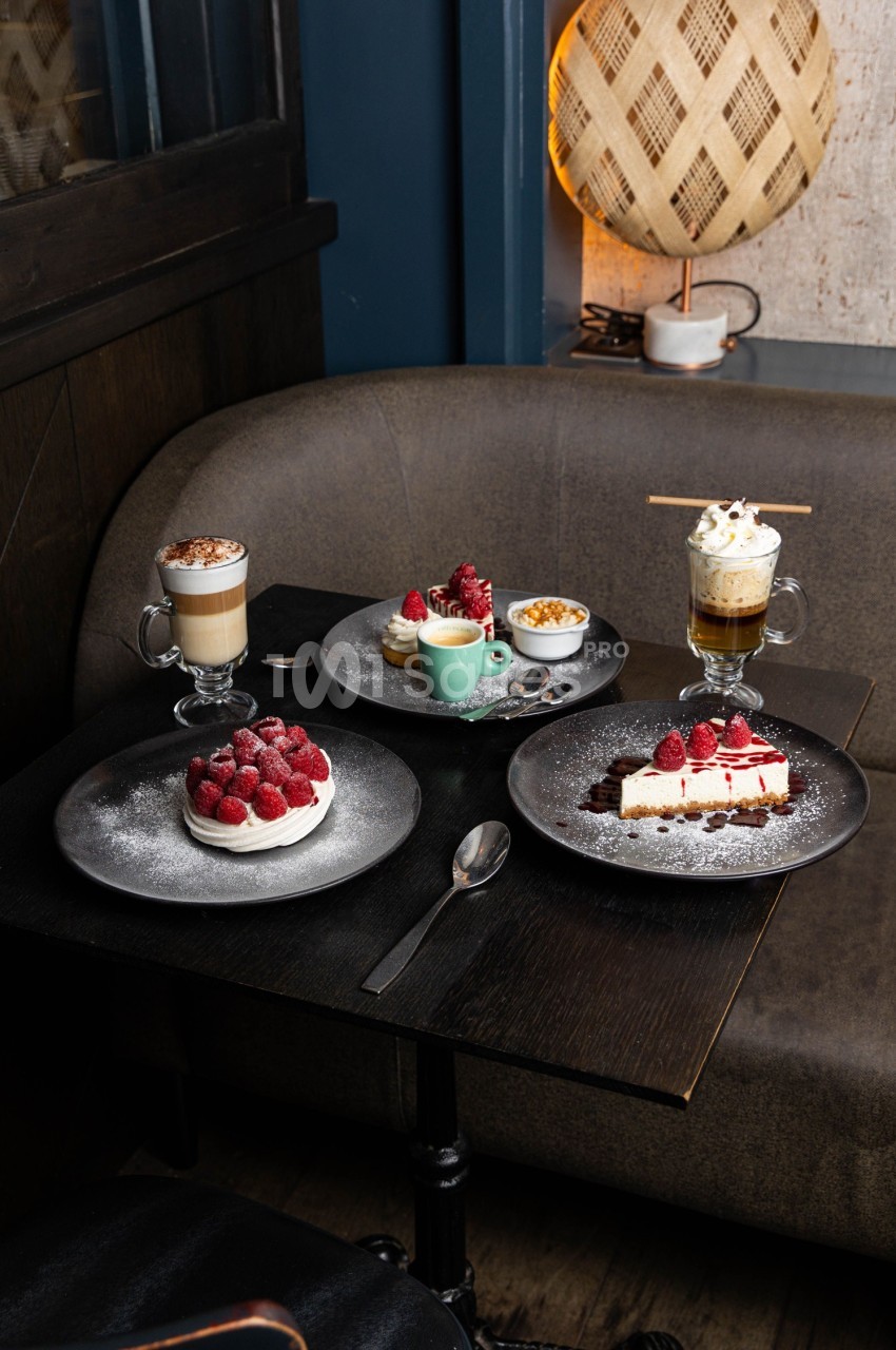 Desserts variés avec framboises, cheesecake, boissons chaudes et garnitures sur une table sombre dans un café cosy.