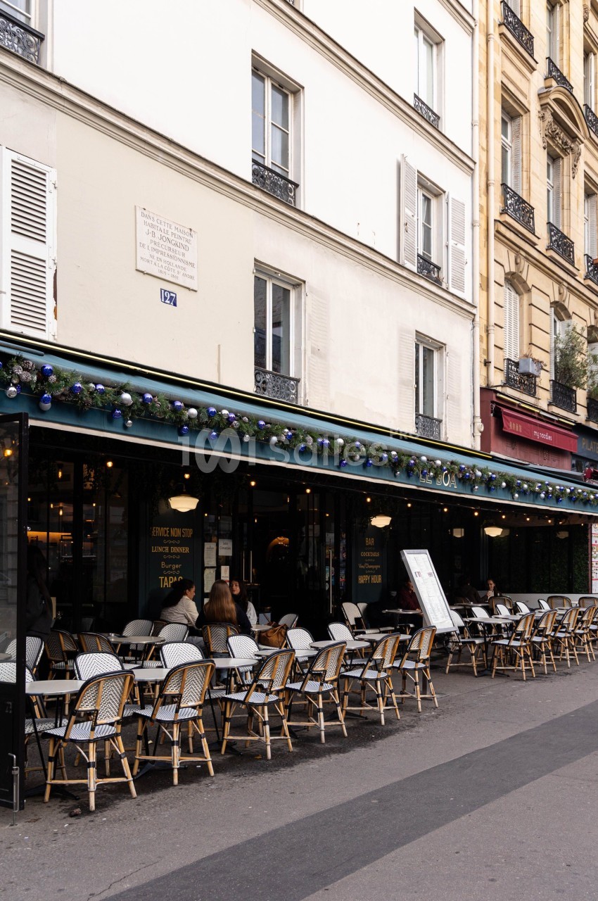 Terrasse de café parisien avec chaises en osier et tables vides, quelques clients assis, façade blanche en arrière-plan.