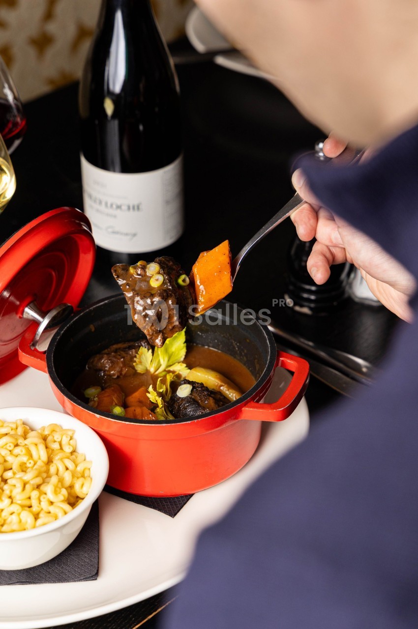 Une personne déguste un plat mijoté servi dans une cocotte rouge, accompagné de pâtes et d'un verre de vin.