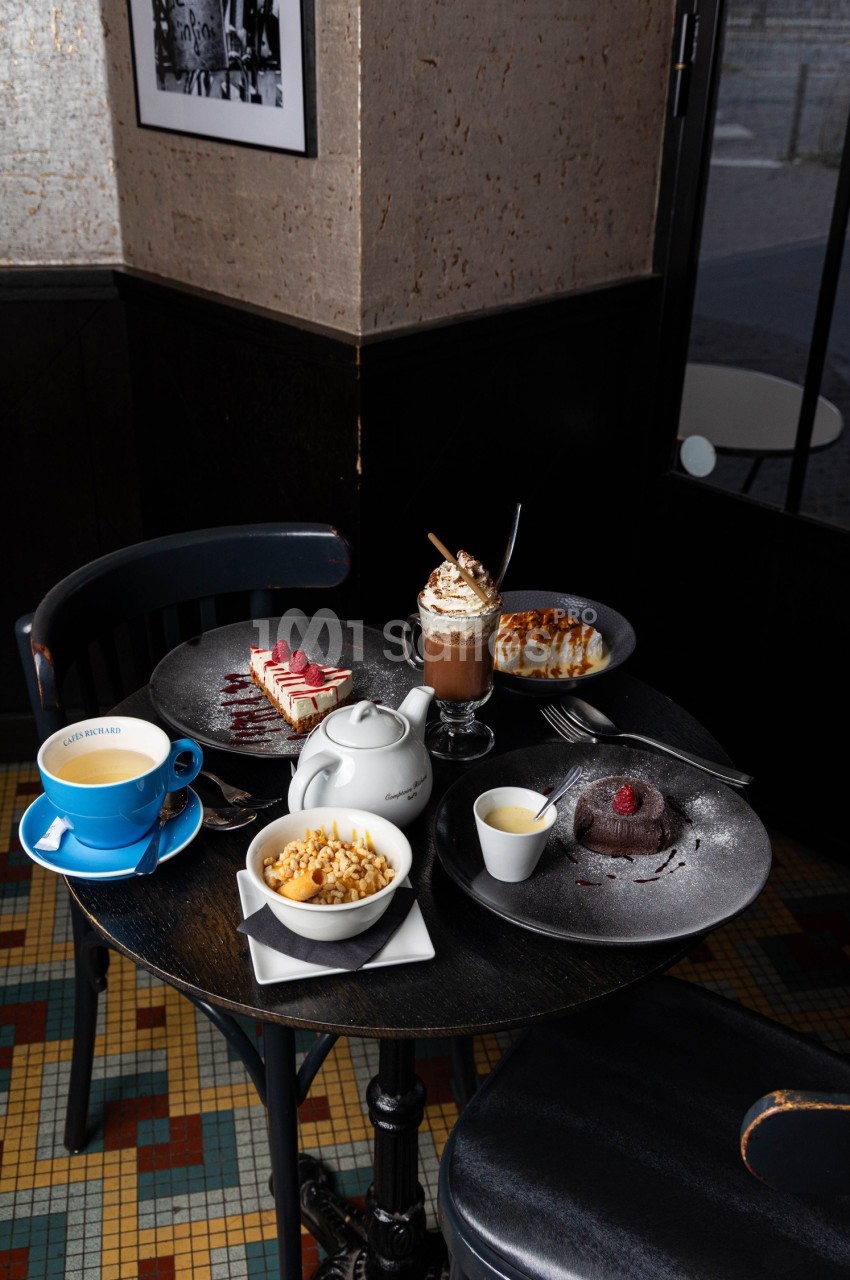 Table ronde avec desserts variés, théière, tasse de café et chocolat chaud, dans un café au sol en mosaïque.